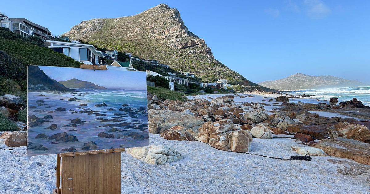 A plein air painting of Misty Cliffs in the foreground, with blue skies, mountain and rocky beach behind it.